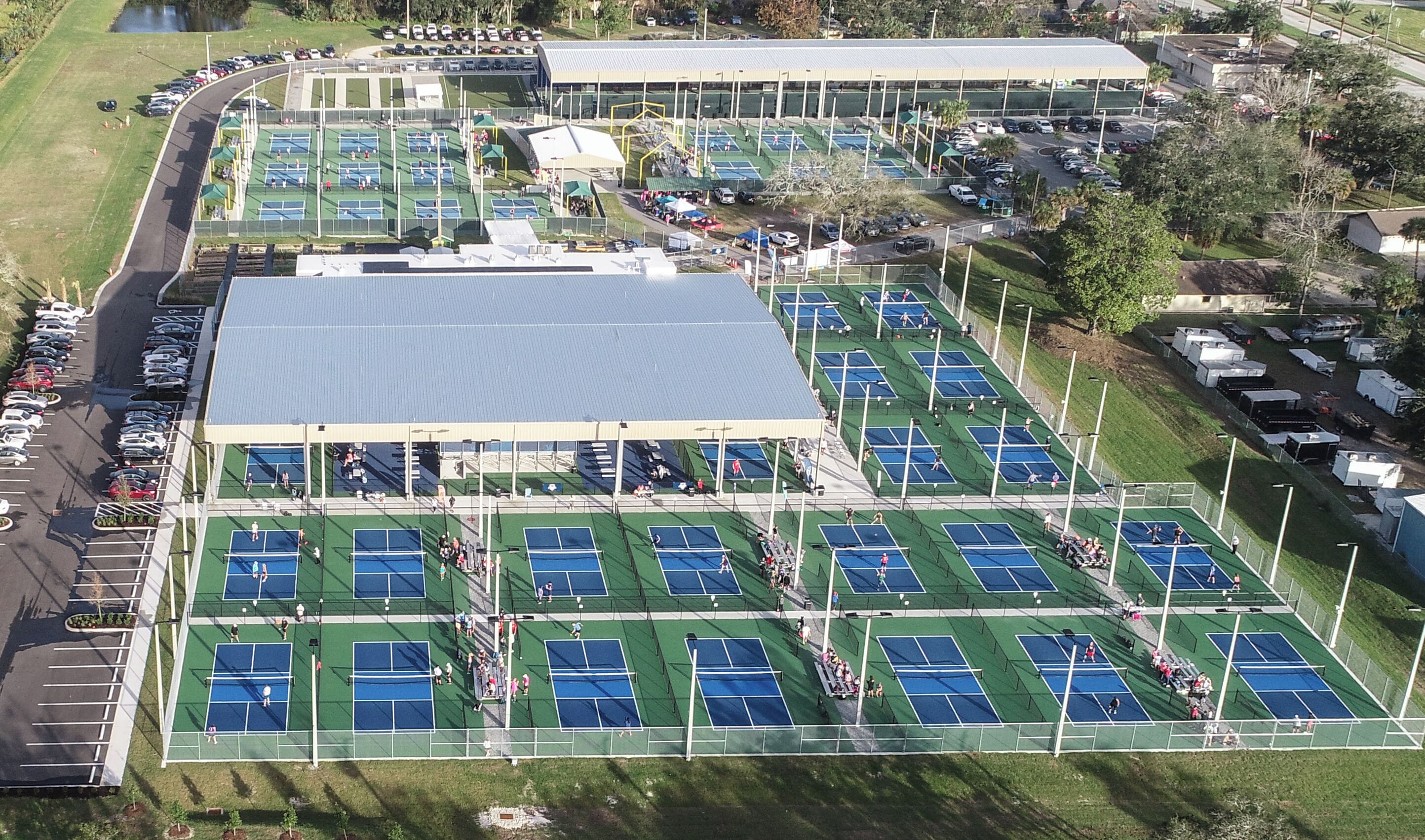 Vista aérea de un complejo deportivo con varias canchas de tenis, una zona de gradas techada y un estacionamiento.