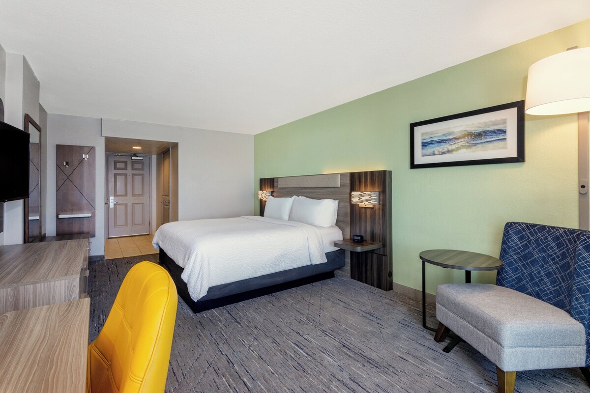 A hotel room with a bed, a desk, a chair, a bedside table, and a lamp.