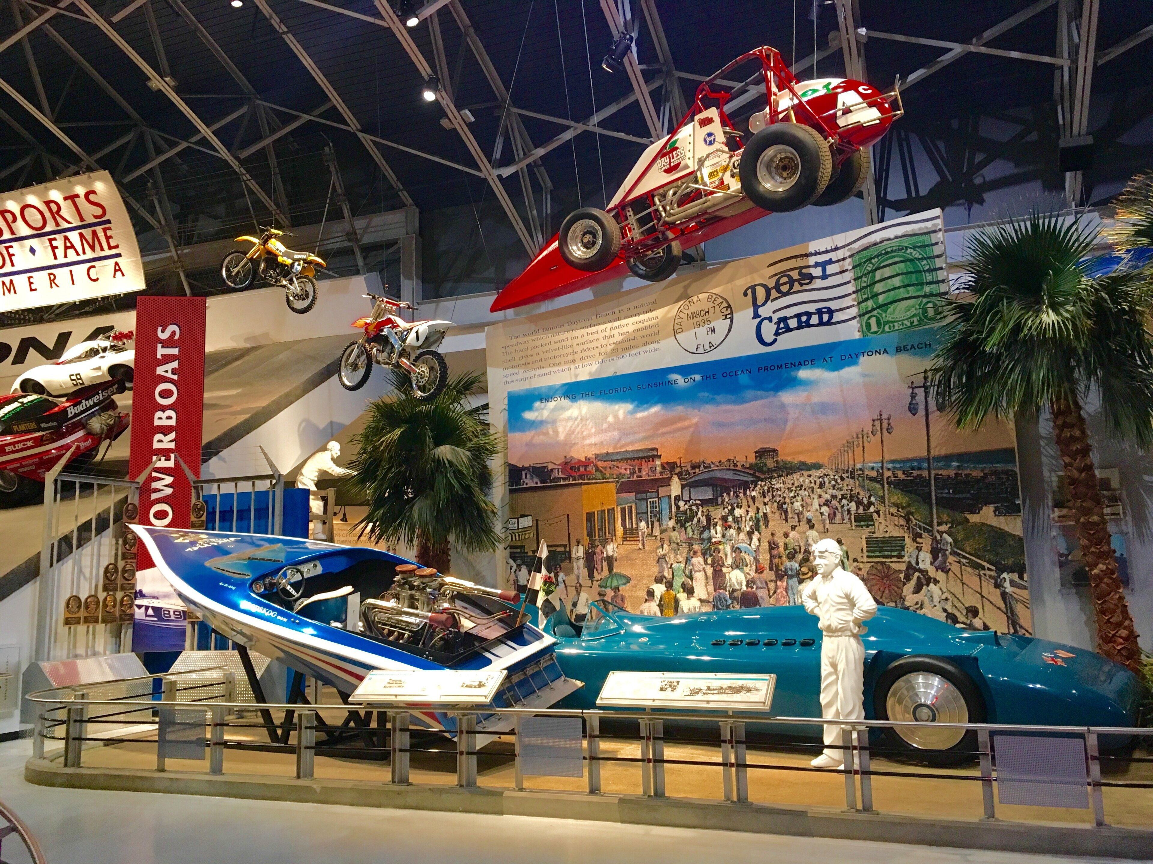 Una exhibición de museo con varios vehículos, incluyendo un barco azul, un auto rojo y una motocicleta.