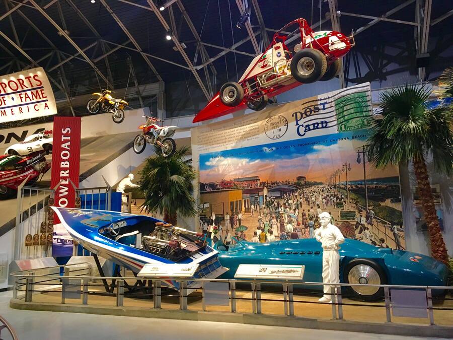 Uma exposição de museu com diversos veículos, incluindo um barco azul, um carro vermelho e uma motocicleta.