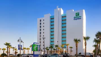 Ein Holiday Inn Express Gebäude mit modernem Design, das ein auffälliges 'H'-Logo zeigt, umgeben von einem Parkplatz mit mehreren Autos und Palmen.