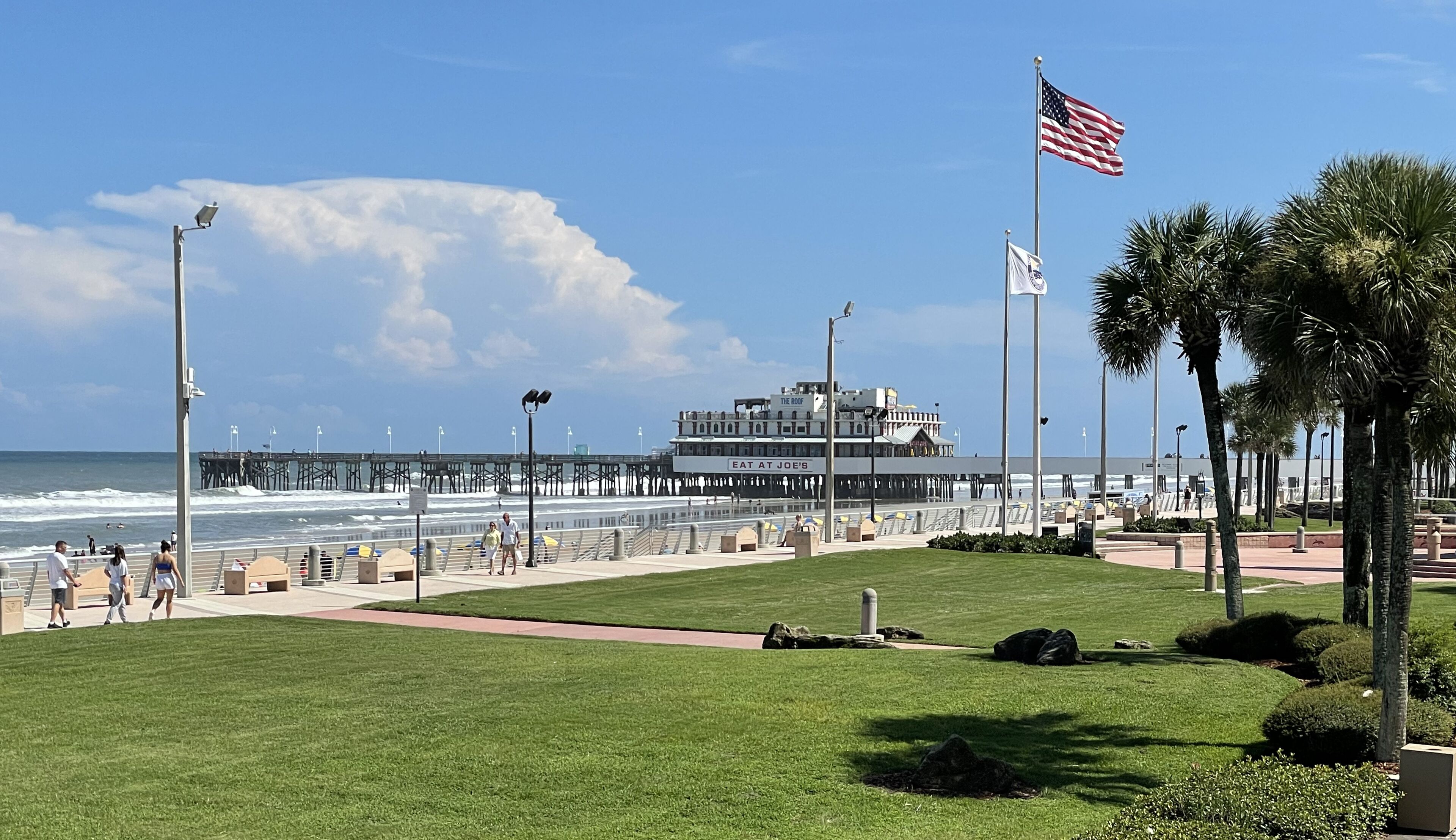 A coastal promenade with a pier, a restaurant named 'The Roof', and a flagpole with the American flag.