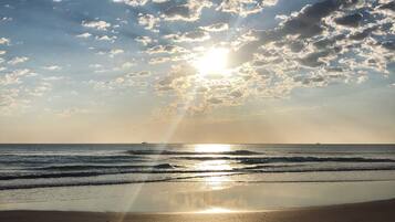 A beach with footprints leading to the water, waves, and a partly cloudy sky with the sun shining.