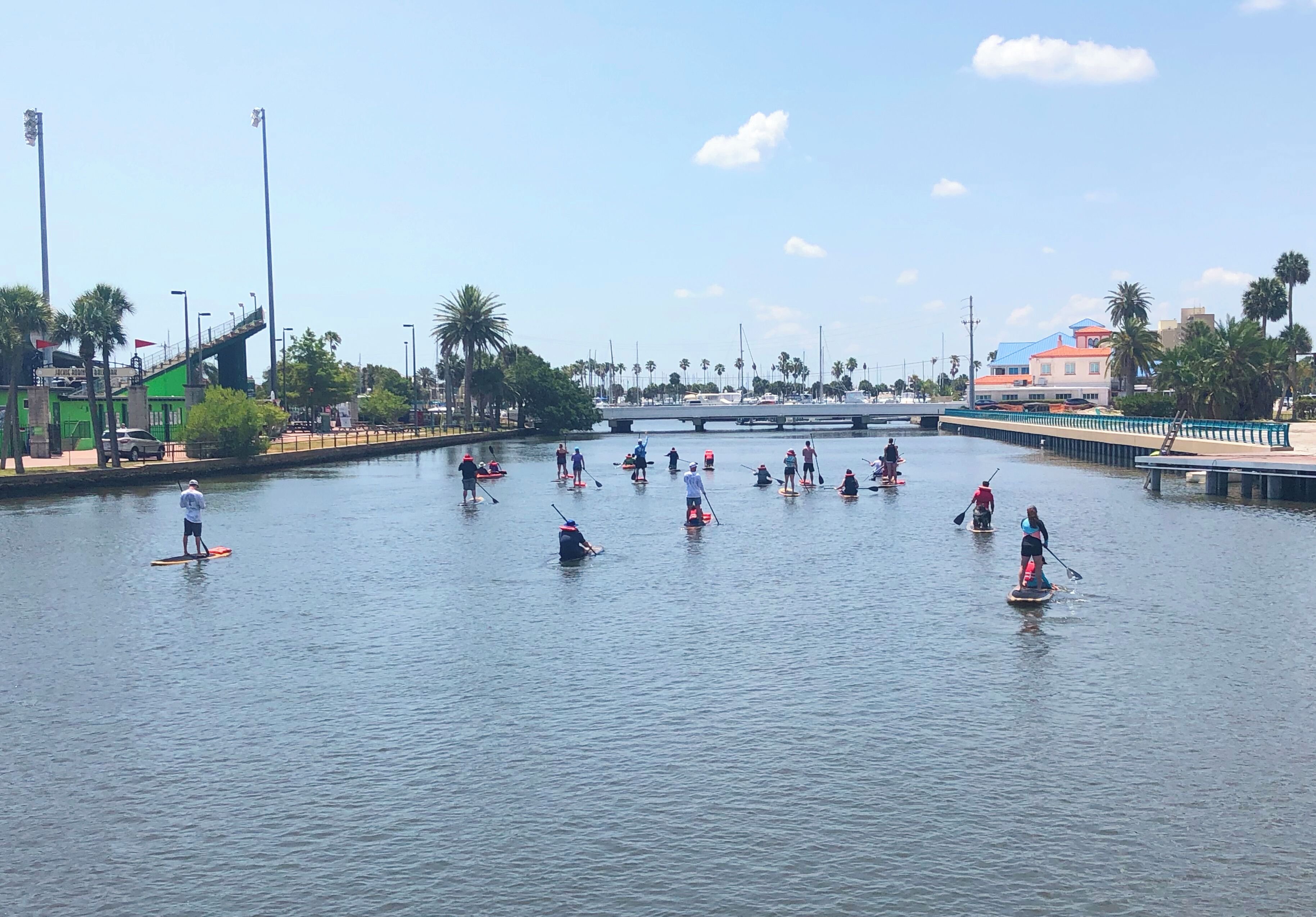 A group of people paddleboarding on a calm water body with a bridge and buildings in the background.