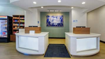 A reception area with two white reception desks, a blue and white wall art piece, a refrigerator stocked with snacks, and a wooden shelf with various items.