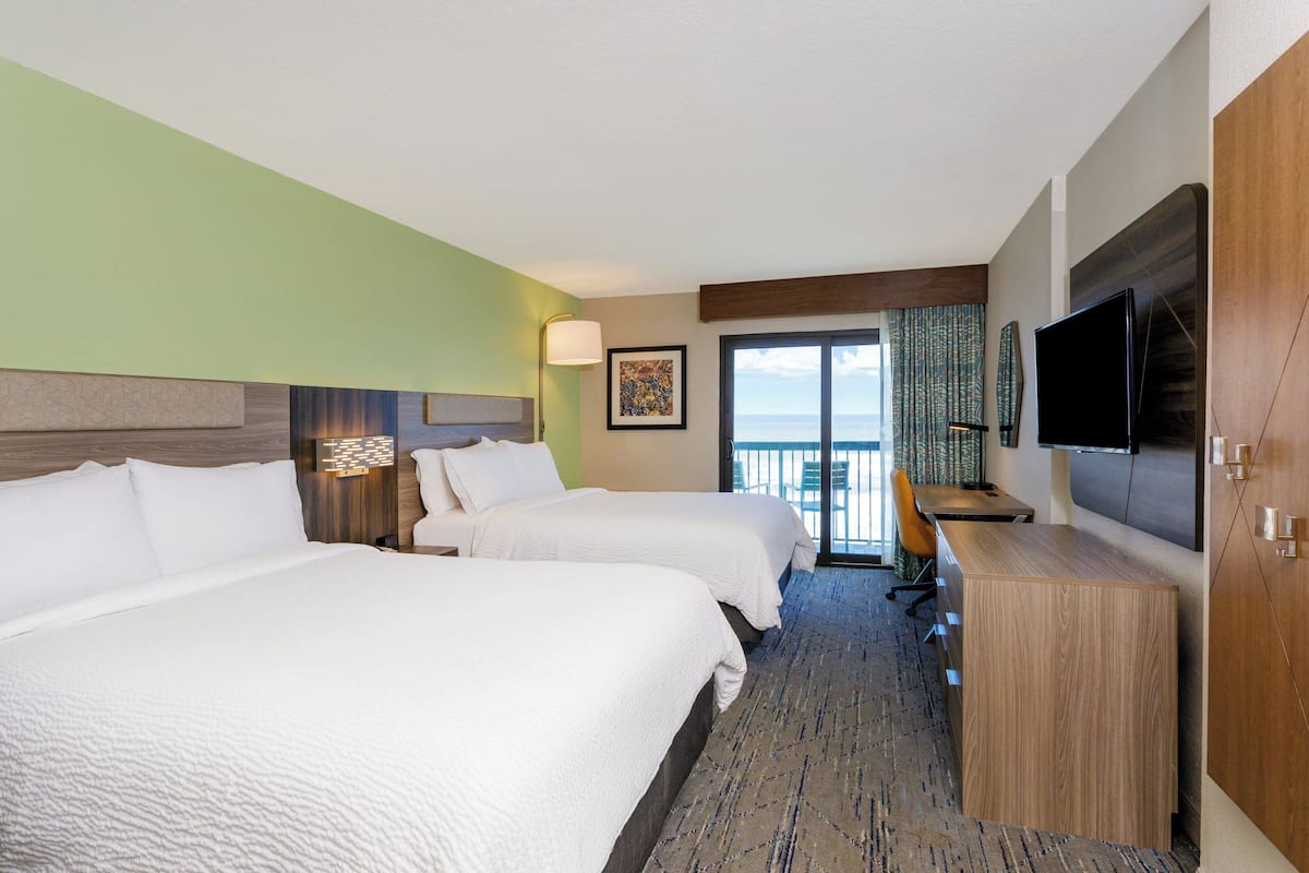 A hotel room with two beds, a desk, a TV, and a view of the ocean.