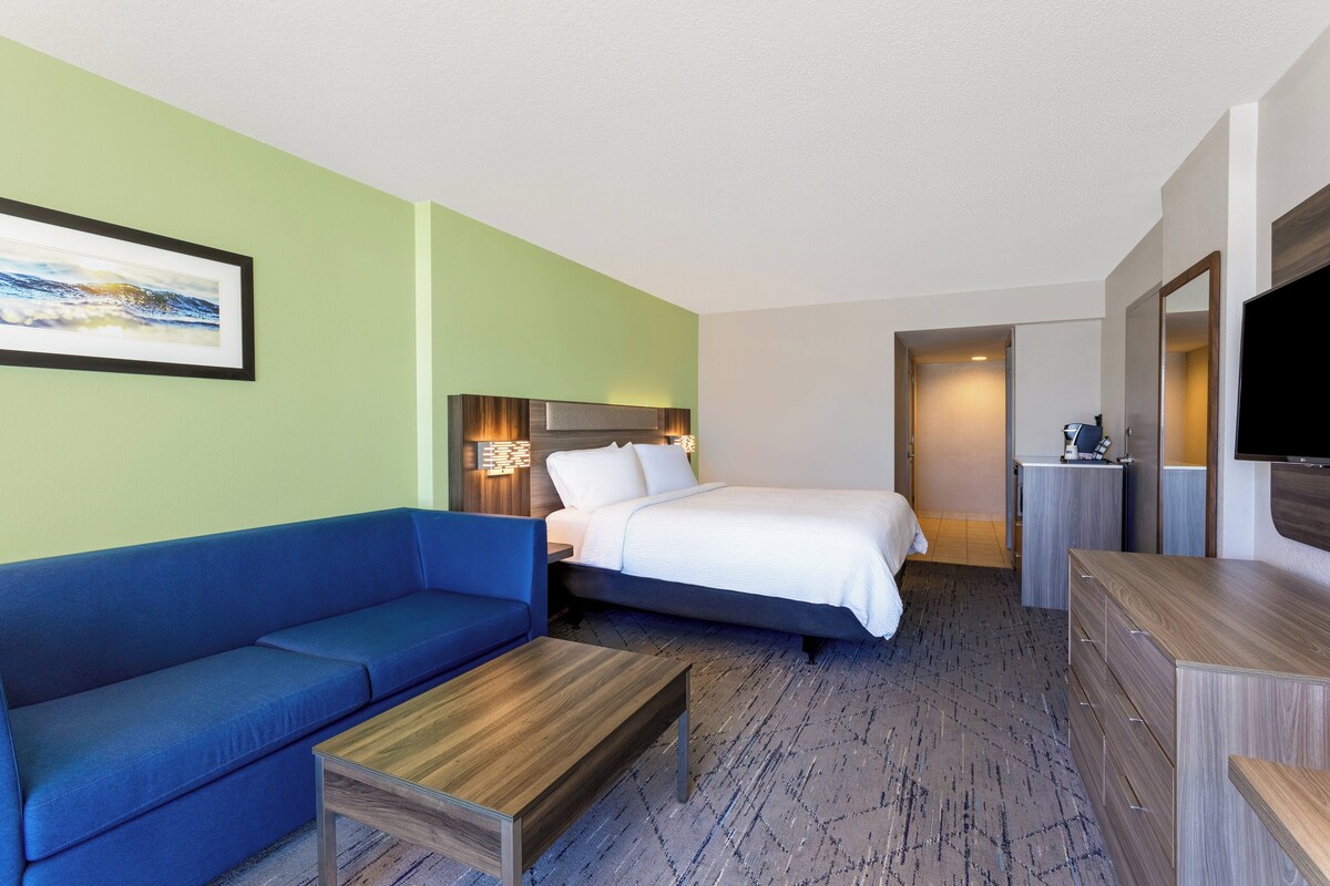 A hotel room with a blue sofa, a bed with white bedding, a wooden coffee table, a dresser, and a flat-screen TV.