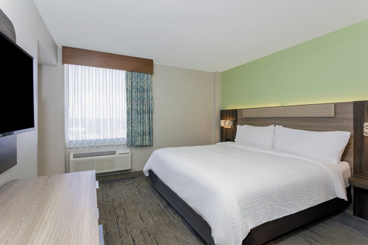 A hotel room with a large bed, a flat-screen TV, a desk, and a window with curtains.