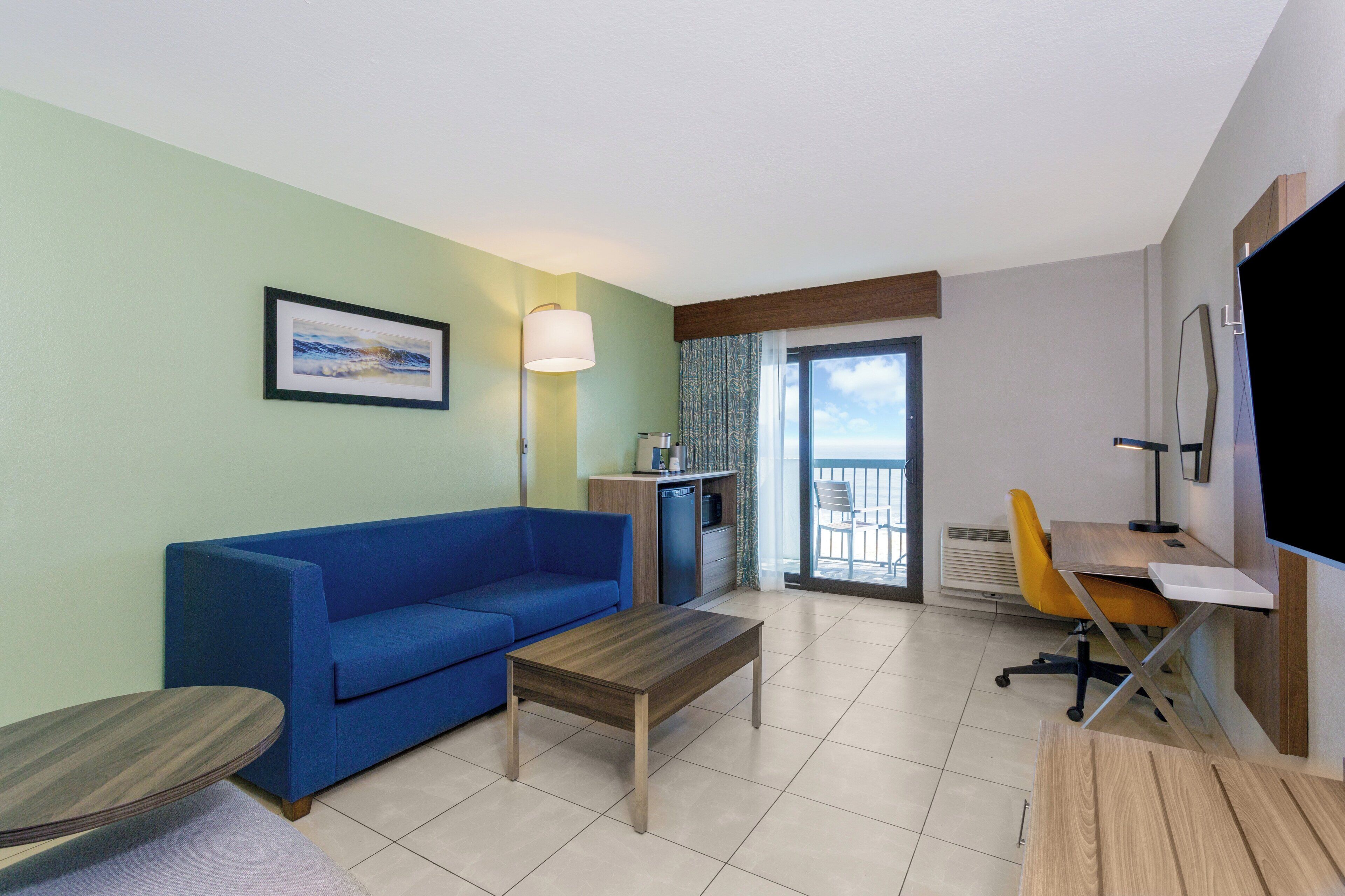 A hotel room with a blue sofa, a wooden coffee table, a desk with a chair, a flat-screen TV, and a framed picture on the wall.