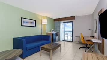 Ein Hotelzimmer mit einem blauen Sofa, einem Holztisch, einem Schreibtisch mit Stuhl, einem Flachbildfernseher und einem gerahmten Bild an der Wand.