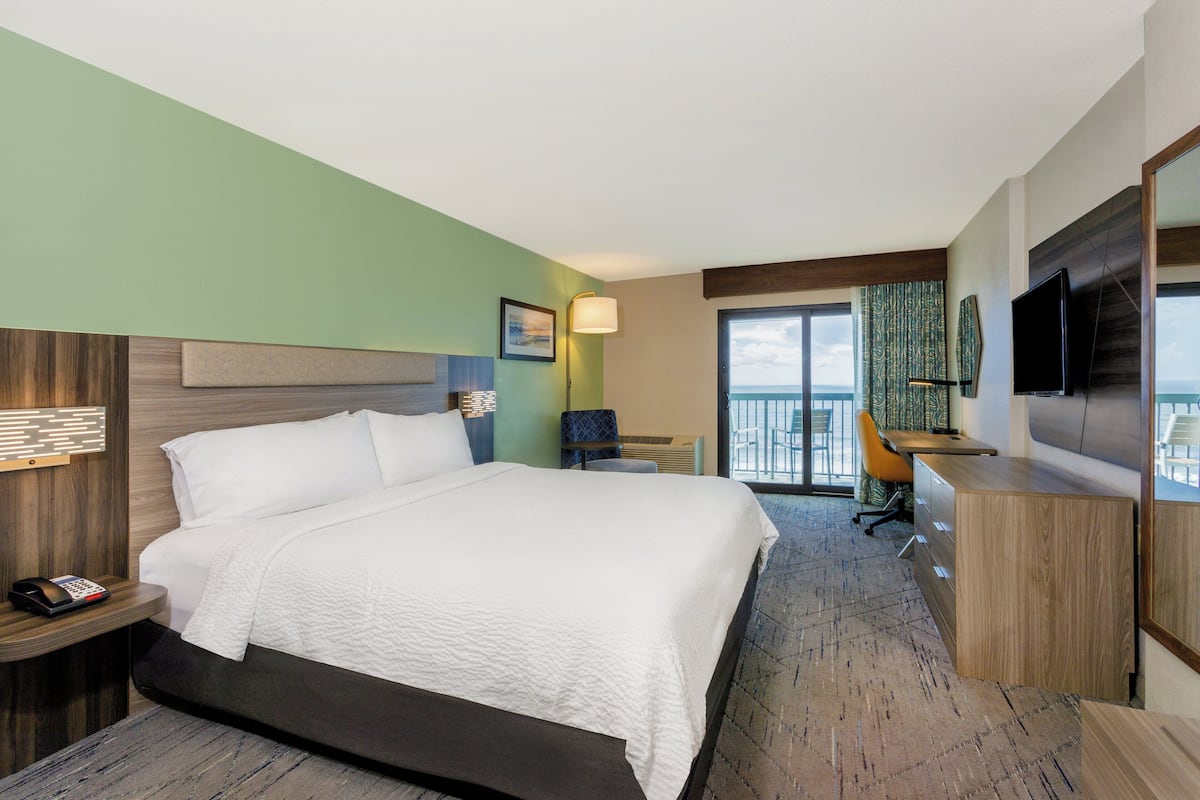A hotel room with a large bed, a desk, a chair, a TV, and a balcony with a view of the ocean.