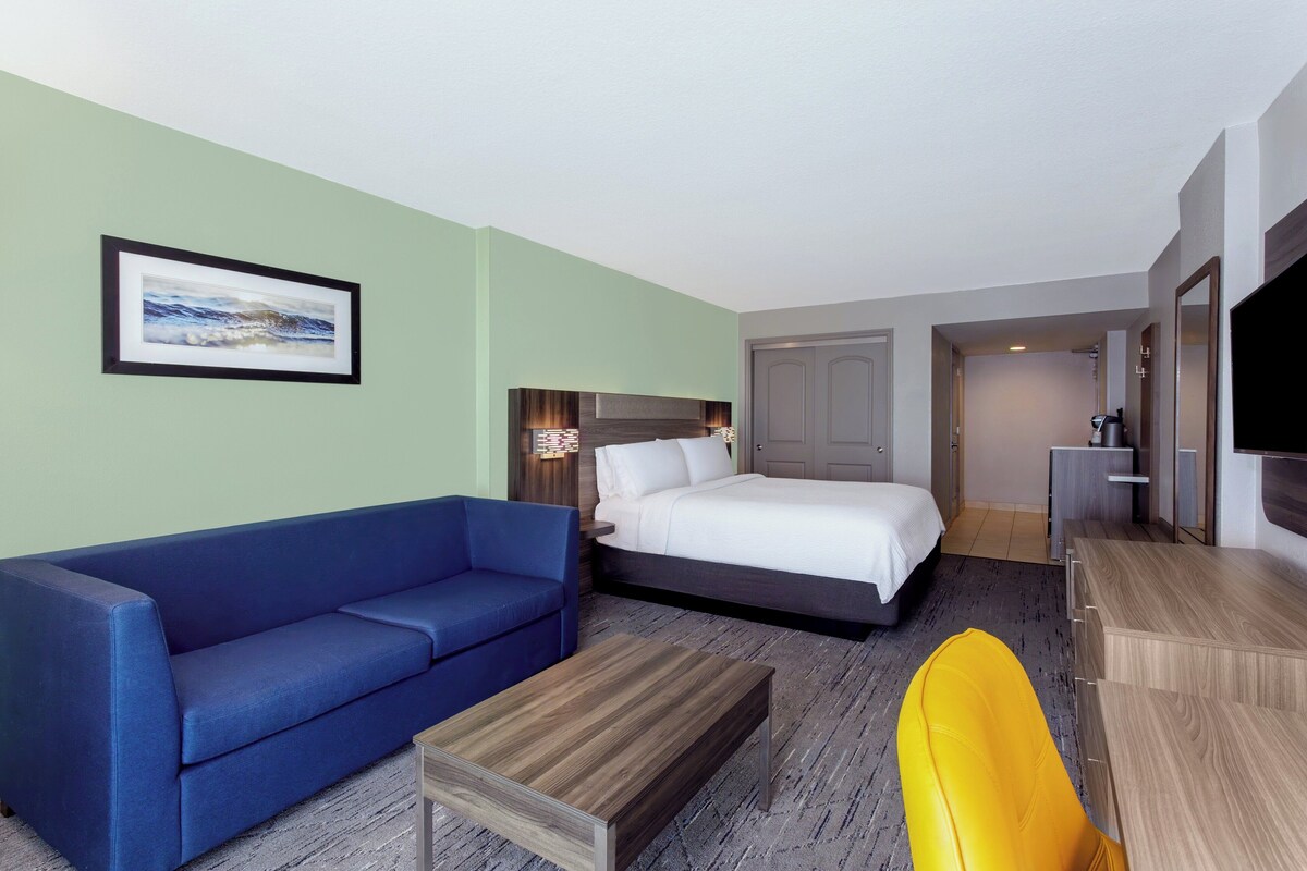 A hotel room with a bed, a blue sofa, a wooden coffee table, a yellow chair, and a flat-screen TV.