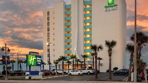 Edifici d'Holiday Inn Express amb un disseny modern, que destaca el logotip prominent 'H', diversos balcons i un aparcament amb diversos cotxes.