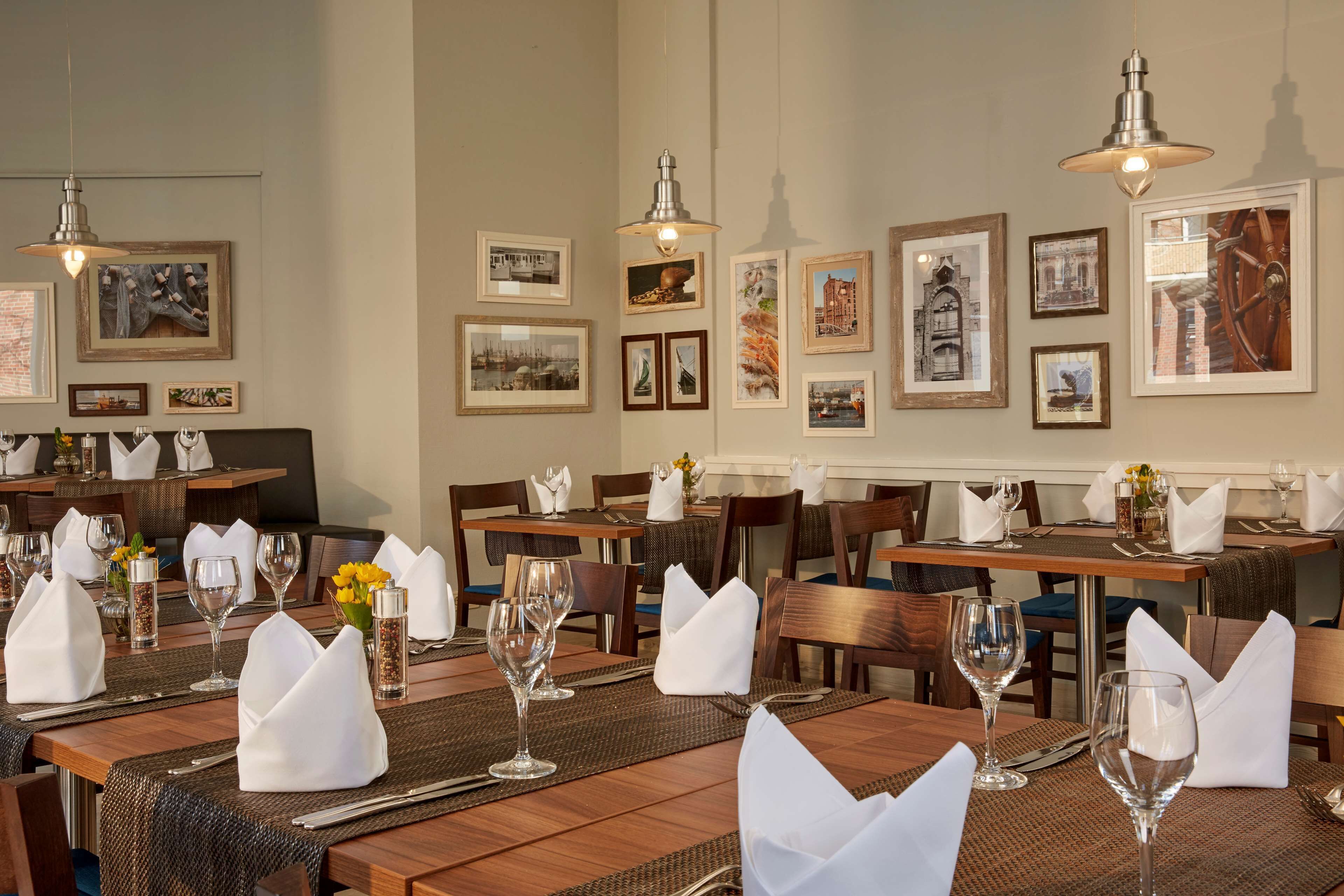 Un restaurant avec des tables en bois, nappées de serviettes blanches, et agrémentées de verres et de couverts. Les murs sont ornés de tableaux encadrés et des lampes sont suspendues au-dessus des tables.