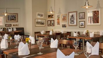 A restaurant with wooden tables set with white napkins, glasses, and cutlery. There are framed pictures on the walls and hanging lights above the tables.