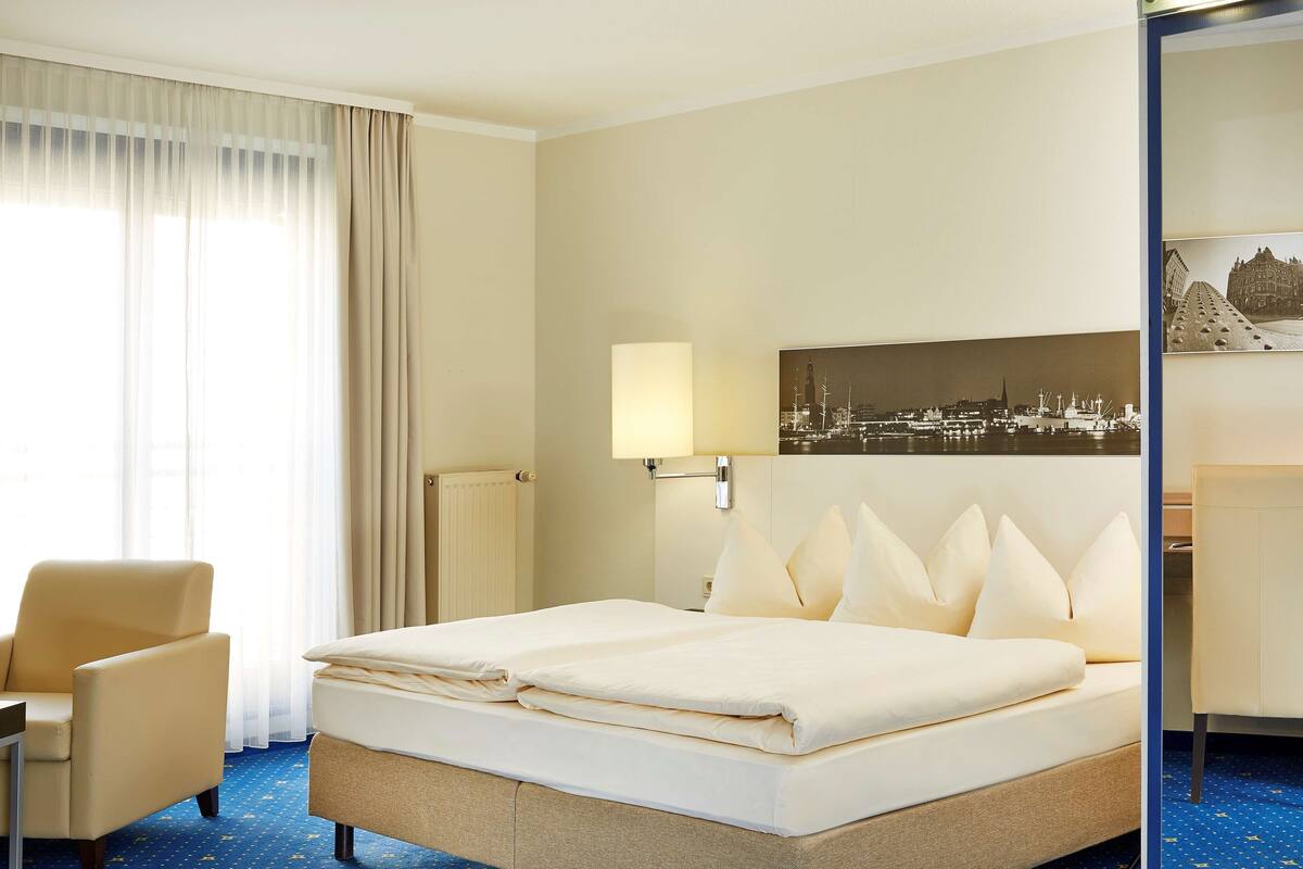 A hotel room with a bed, a chair, a lamp, and a cityscape painting on the wall.