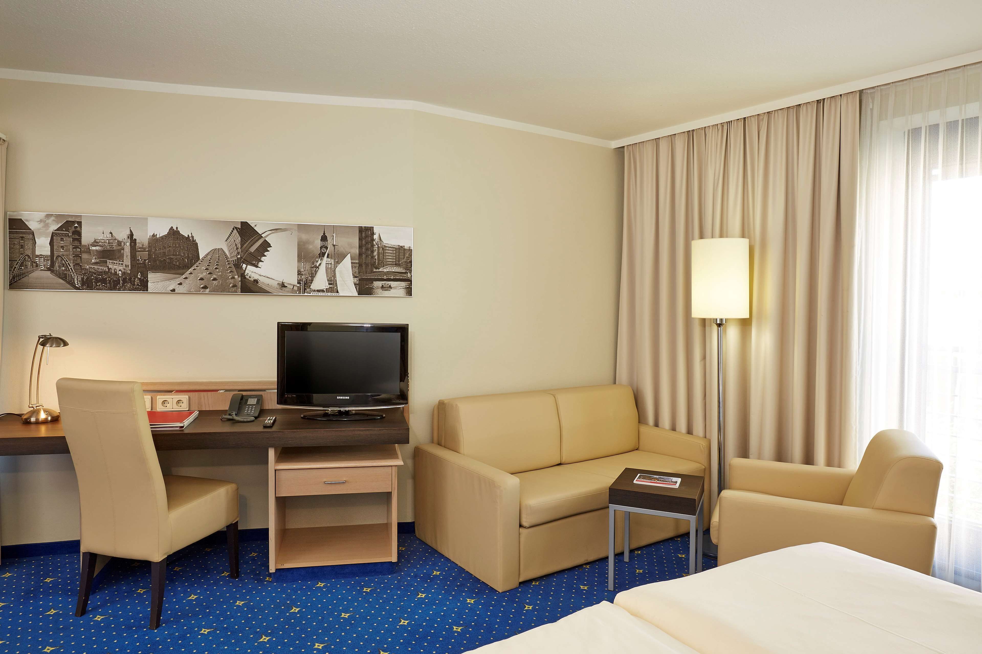 A hotel room with a bed, a desk with a computer, a sofa, a chair, and a TV.