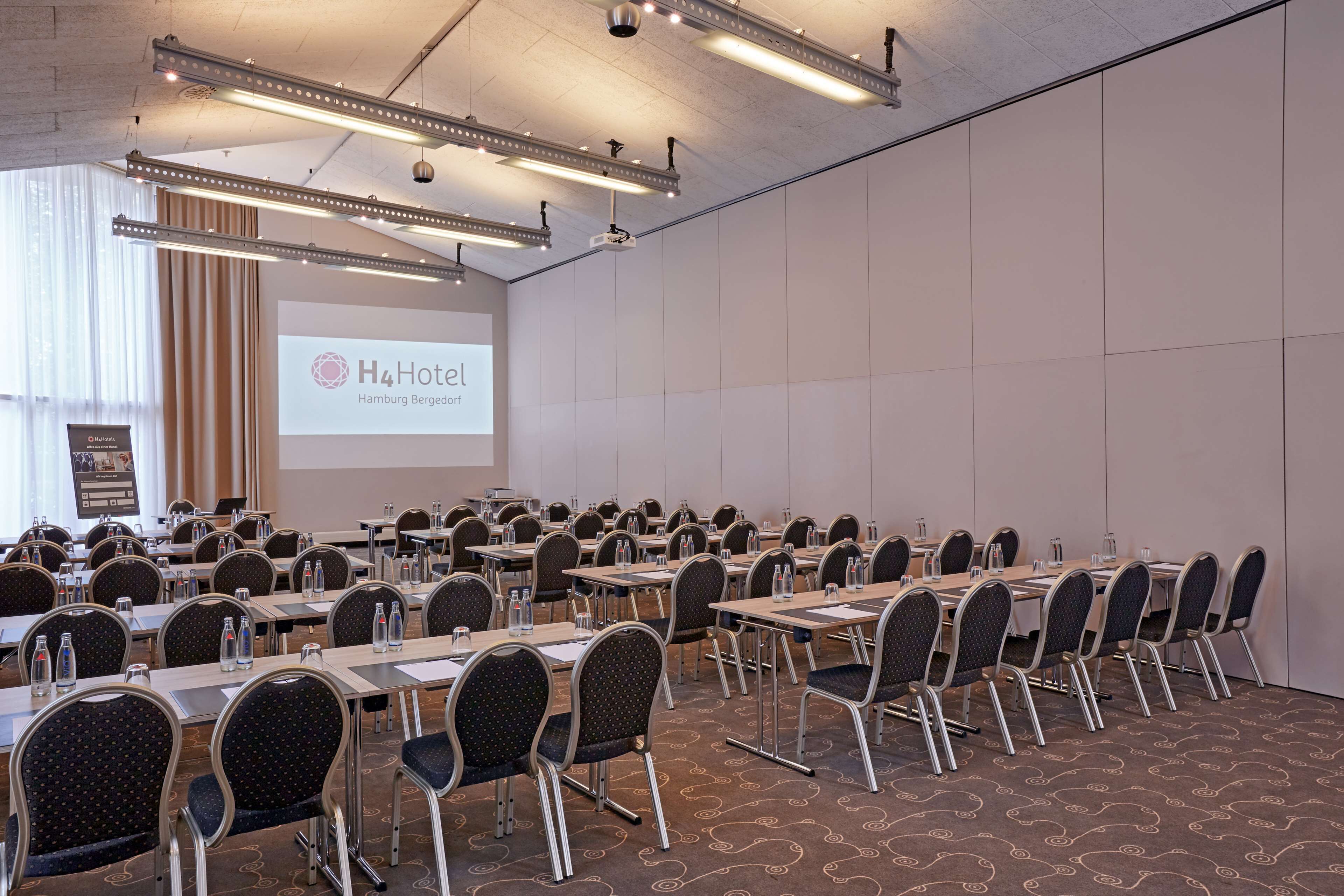 Un salón de conferencias con varias mesas y sillas, una pantalla de proyector mostrando el logotipo de H4Hotel Hamburg Bergedorf y un gran ventanal con cortinas.