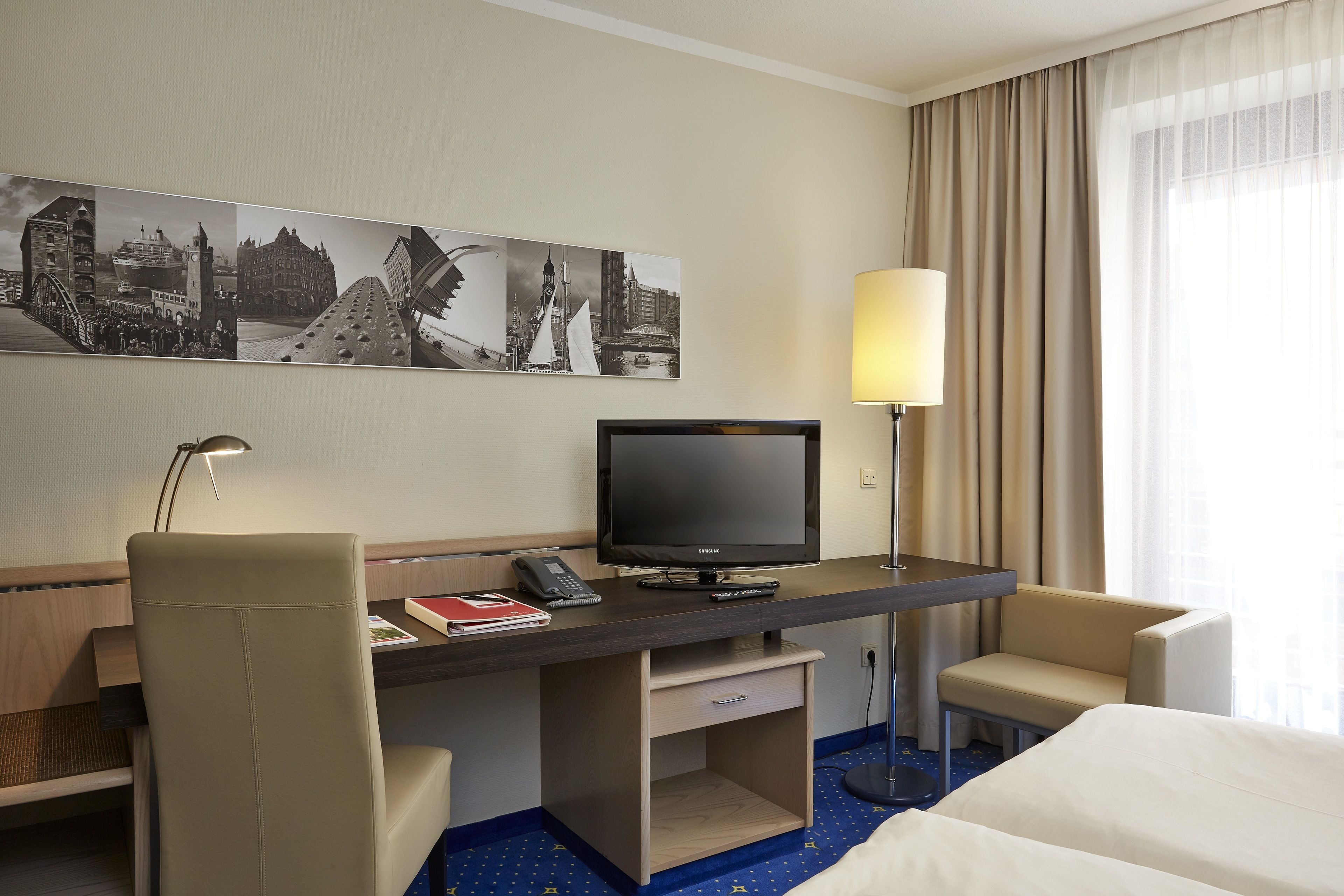 A hotel room with a bed, a desk with a television, a chair, a lamp, and a wall with a cityscape artwork.