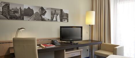 A hotel room with a bed, a desk with a television, a chair, a lamp, and a wall with a cityscape artwork.