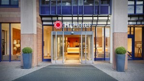 Entrada de hotel con puerta de cristal, un logotipo rojo y dos plantas en macetas.
