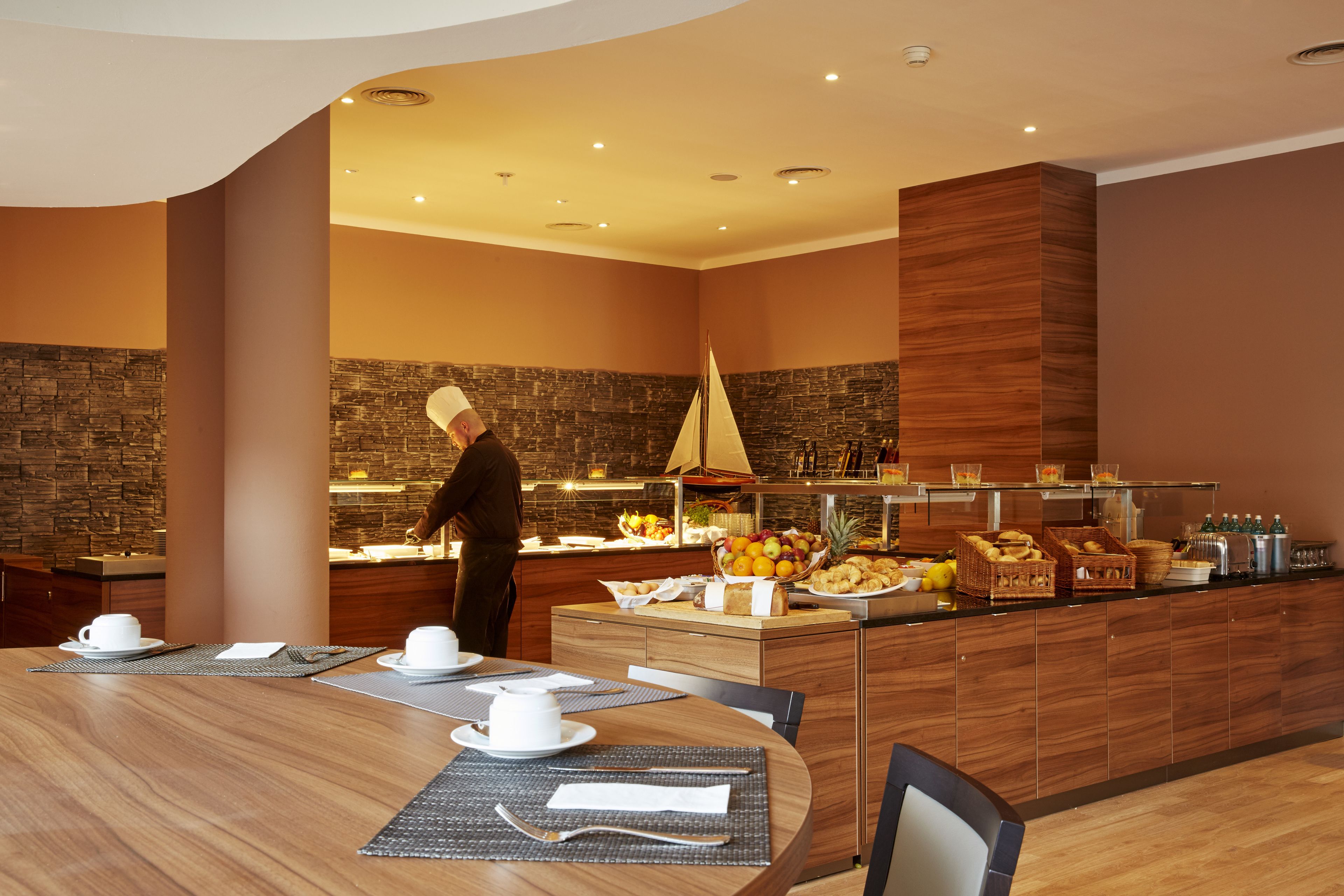 Een modern restaurantgedeelte van een hotel, met een kok die eten bereidt, een buffet met fruit en snacks, en een houten eettafel gedekt voor twee personen.