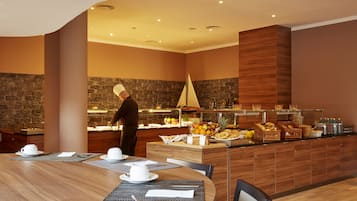Een modern restaurantgedeelte van een hotel, met een kok die eten bereidt, een buffet met fruit en snacks, en een houten eettafel gedekt voor twee personen.