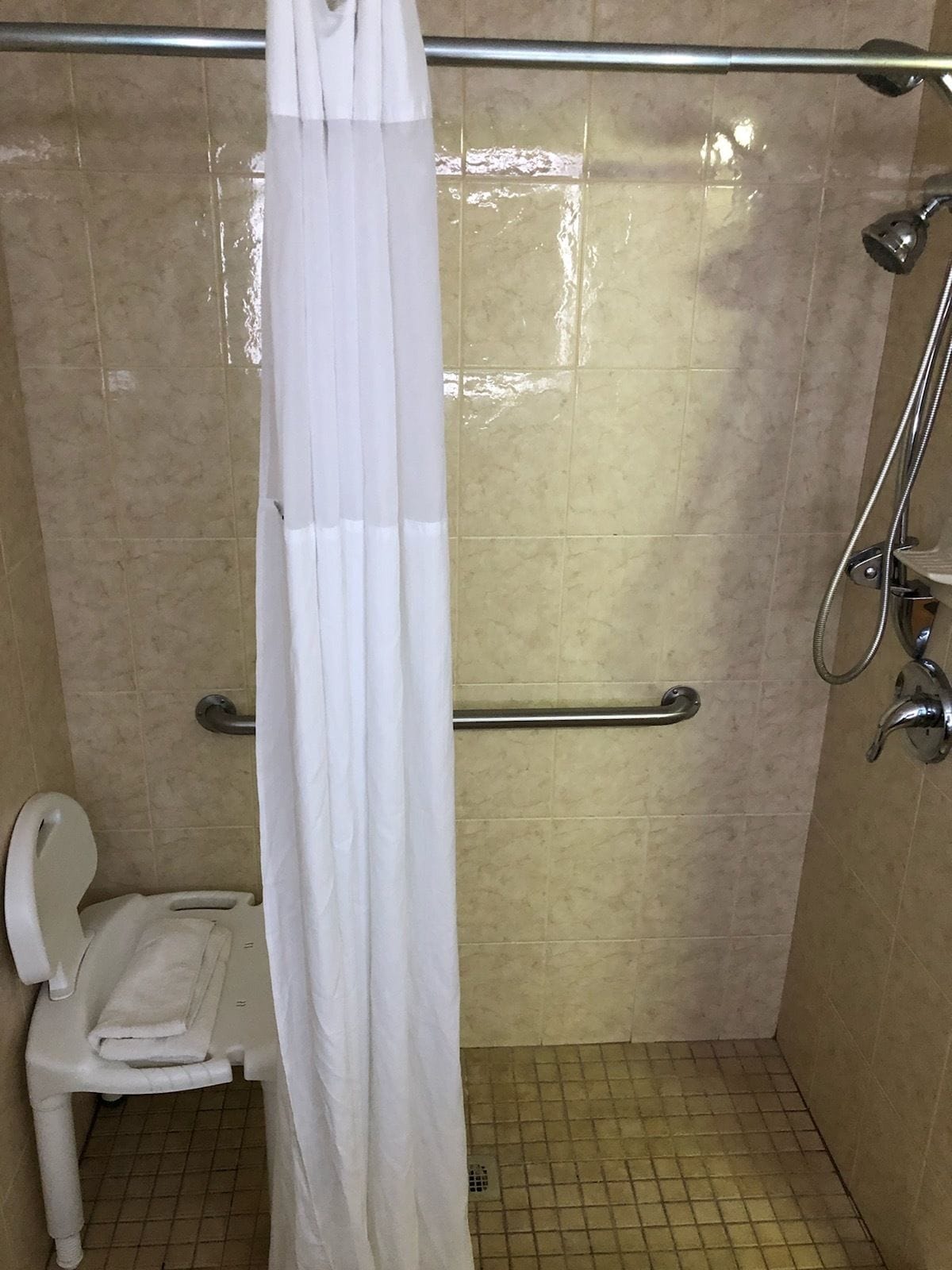A shower area with a grab bar, a white chair, and a white shower curtain.
