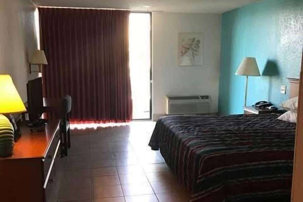 Une chambre d’hôtel avec un lit, un bureau doté d’une lampe, une chaise, une fenêtre avec des rideaux et un mur orné d’un tableau.