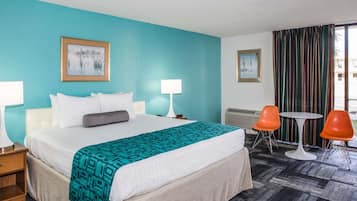 Habitación de hotel con una cama grande, dos mesitas de noche con lámparas, una pequeña mesa redonda con dos sillas naranjas y una ventana con cortinas.