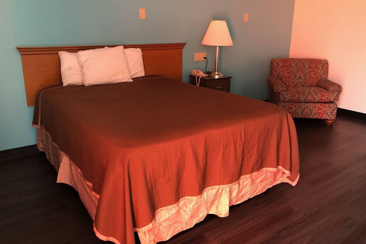 Un lit bien fait, recouvert d’une couverture marron et agrémenté d’oreillers blancs, une lampe de chevet, un téléphone et un fauteuil à motifs dans une pièce aux murs bleu clair et au plancher en bois foncé.