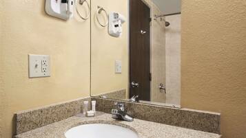 A bathroom with a granite countertop sink, a mirror, a hairdryer, and a towel dispenser.