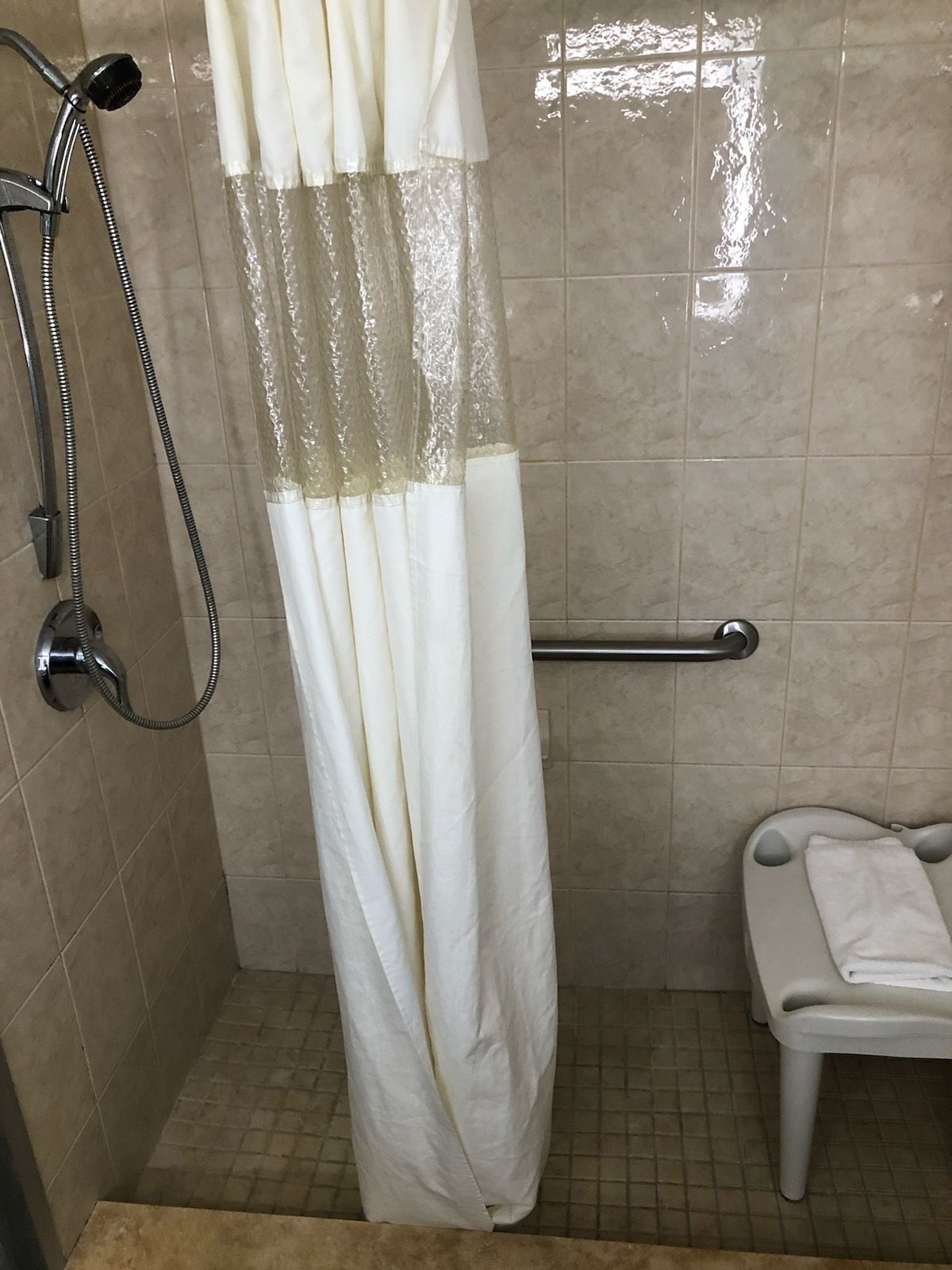 A shower area with a white shower curtain, a handheld showerhead, a grab bar, and a small white bench with a towel.