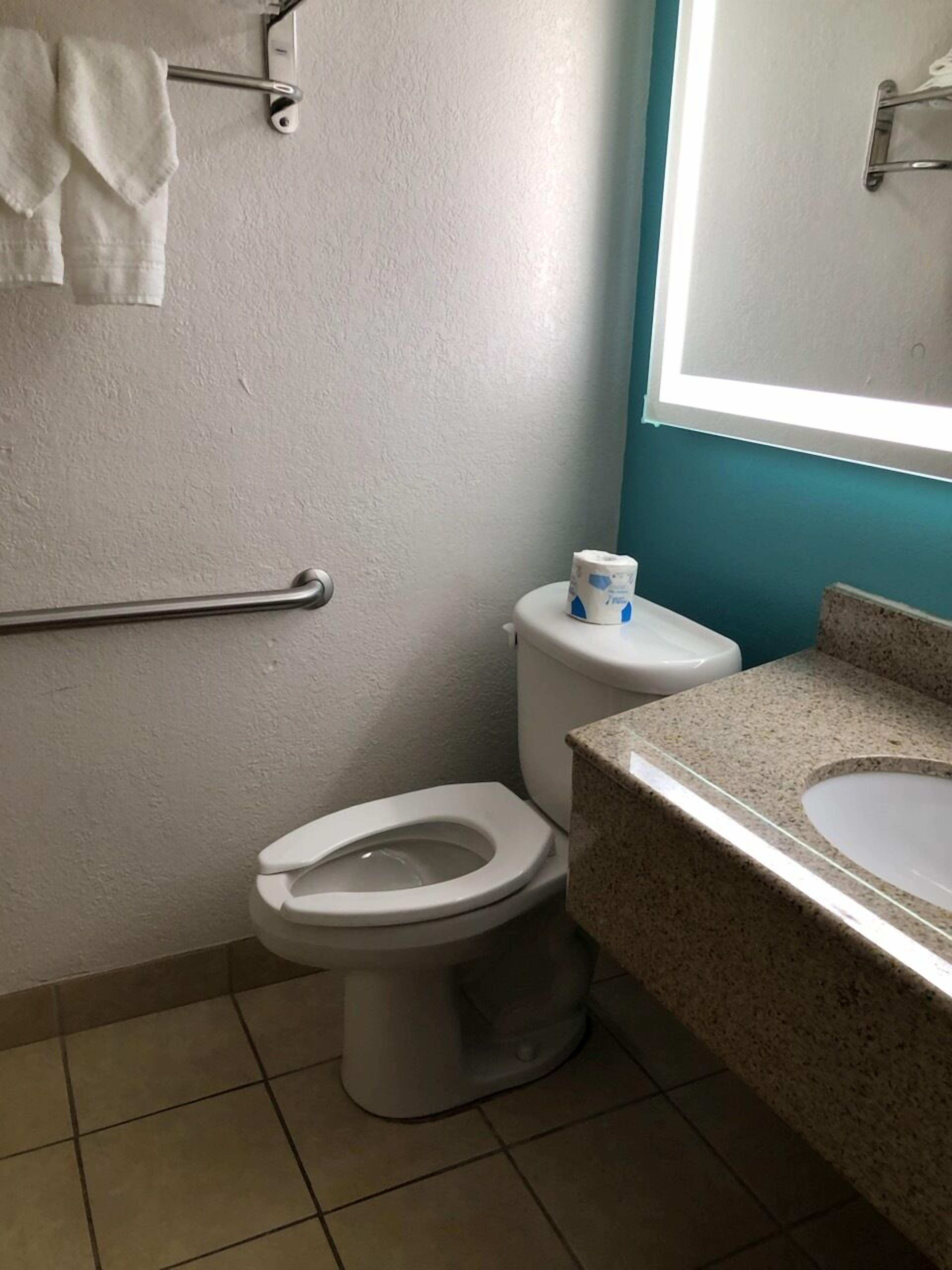 Un baño con inodoro blanco, barra de apoyo, lavamanos con encimera de granito y dispensador de papel higiénico.