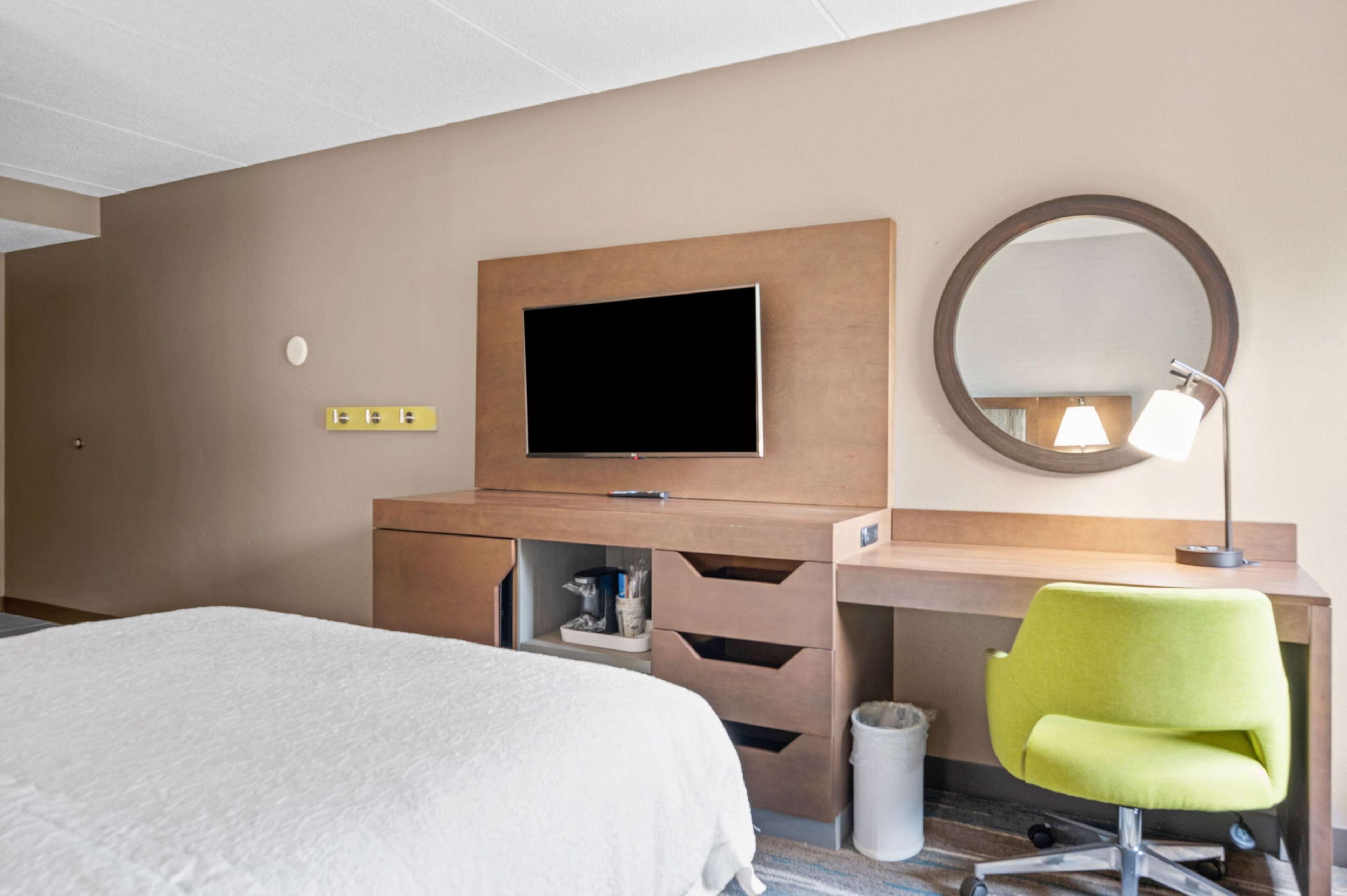 A hotel room with a bed, a desk with a chair, a television, a mirror, and a lamp.