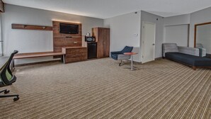Habitación de hotel con televisor de pantalla plana en la pared, microondas, una mesita con una silla, sofá y un banco.