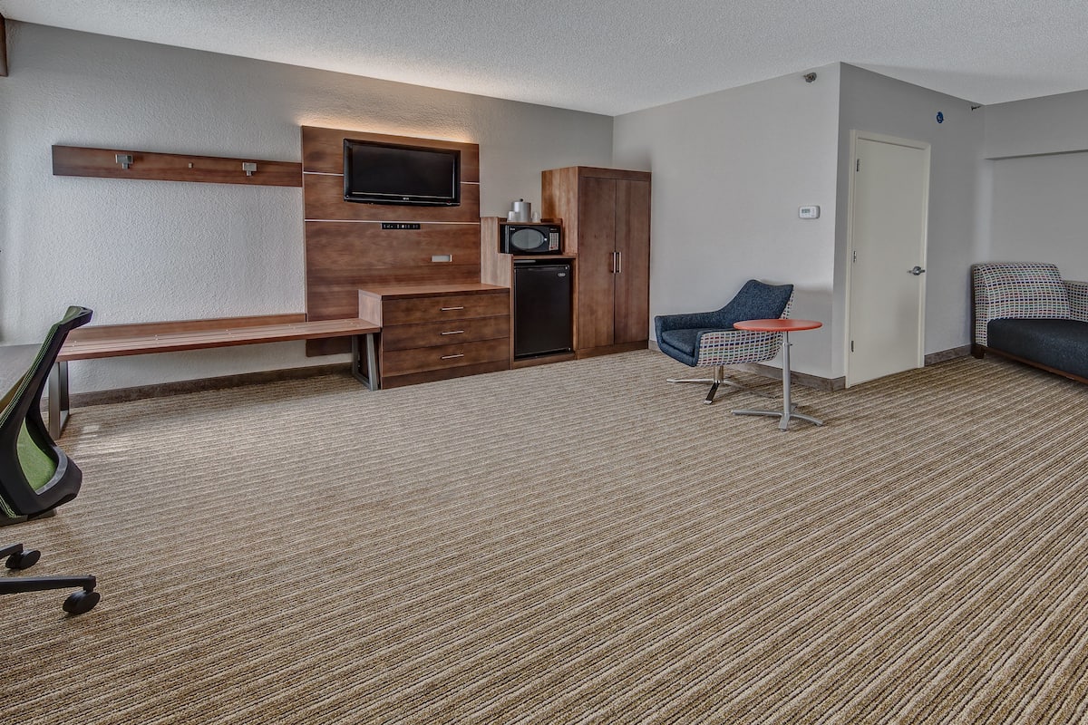 A hotel room with a flat-screen TV mounted on the wall, a microwave, a small table with a chair, a sofa, and a bench.