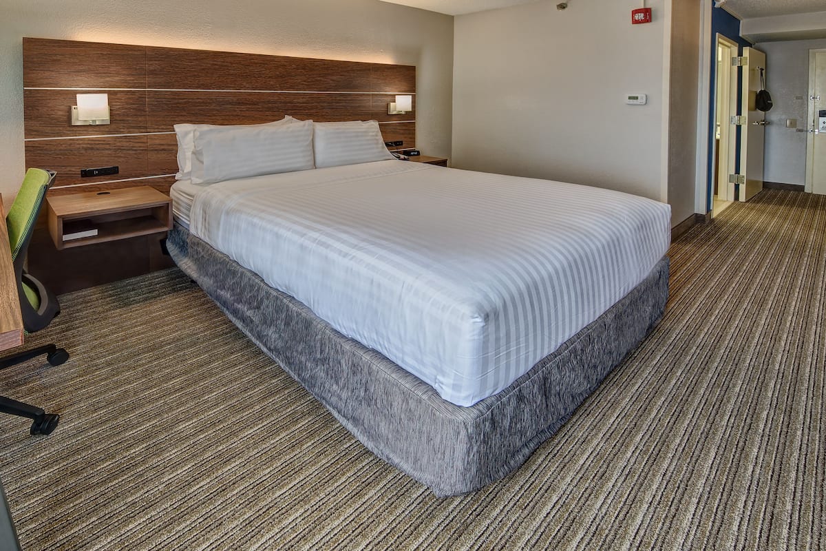 A hotel room with a large bed, a desk, and a chair. There are two wall-mounted lights above the bed and a thermostat on the wall.