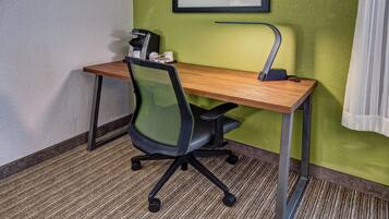 A hotel room with a wooden desk, a black office chair, a coffee machine, and a green textured wall.