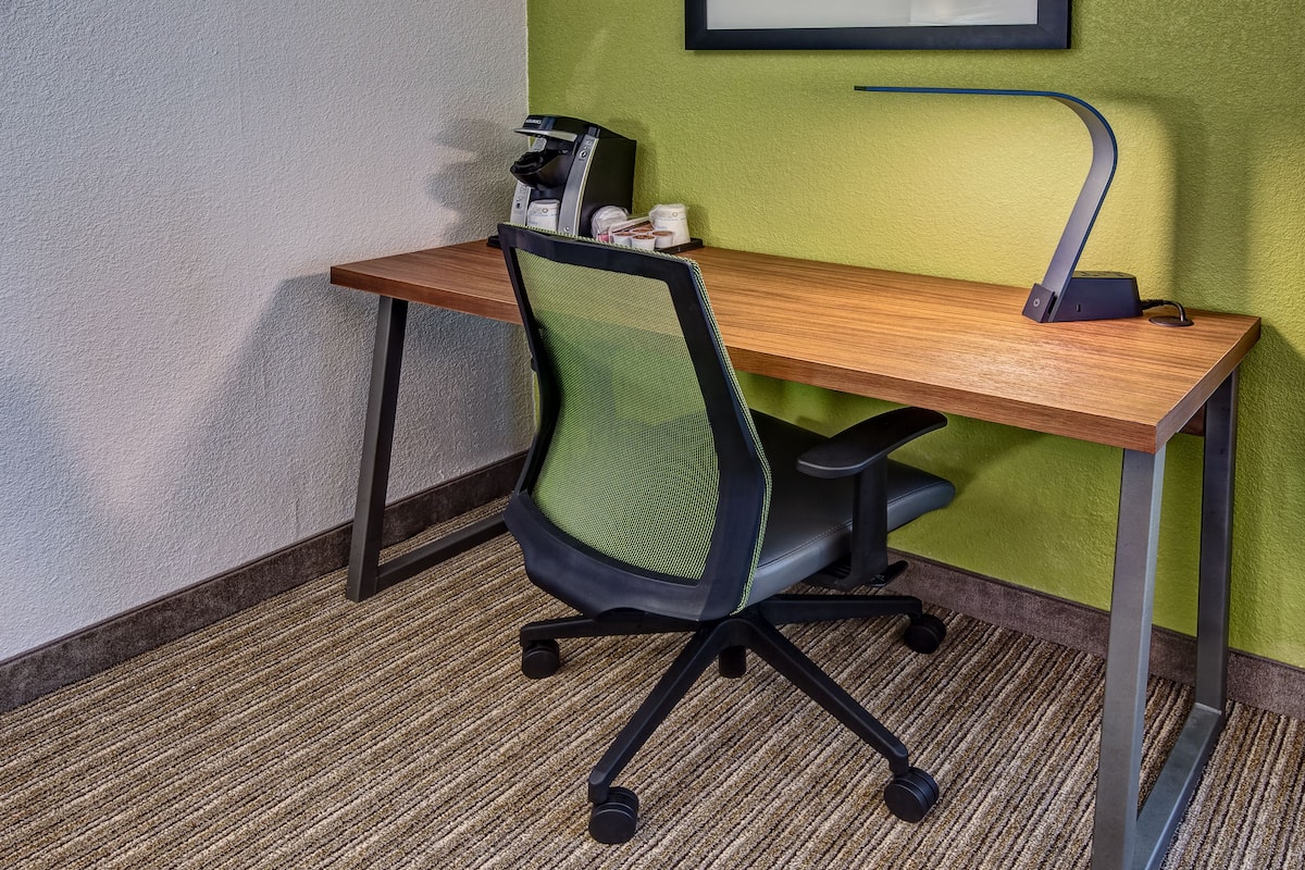 A hotel room with a wooden desk, a black office chair, a coffee machine, and a green wall.