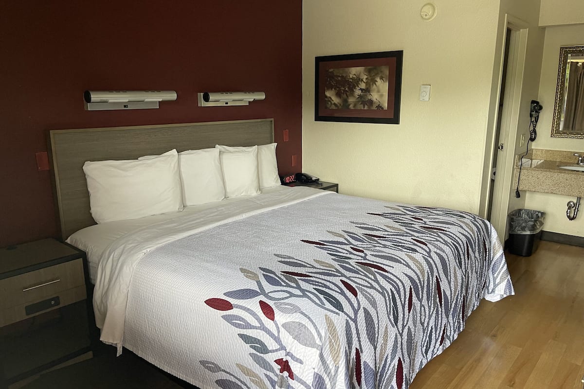 A hotel room with a bed, bedside tables, a nightstand, a framed picture on the wall, and a bathroom visible through an open door.