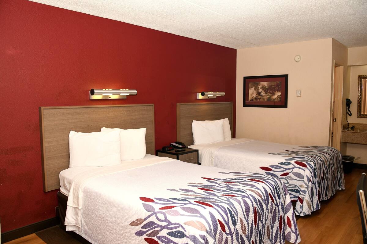 A hotel room with two beds, each with a headboard, a bedside table, and a lamp.