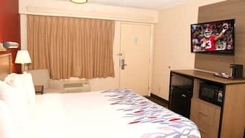 A hotel room with a bed, a television mounted on the wall, a desk with a microwave, and a door with a curtain.