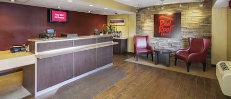 A hotel reception area with a digital display, a reception desk, a seating area with chairs, and a Red Roof Inn sign on the wall.