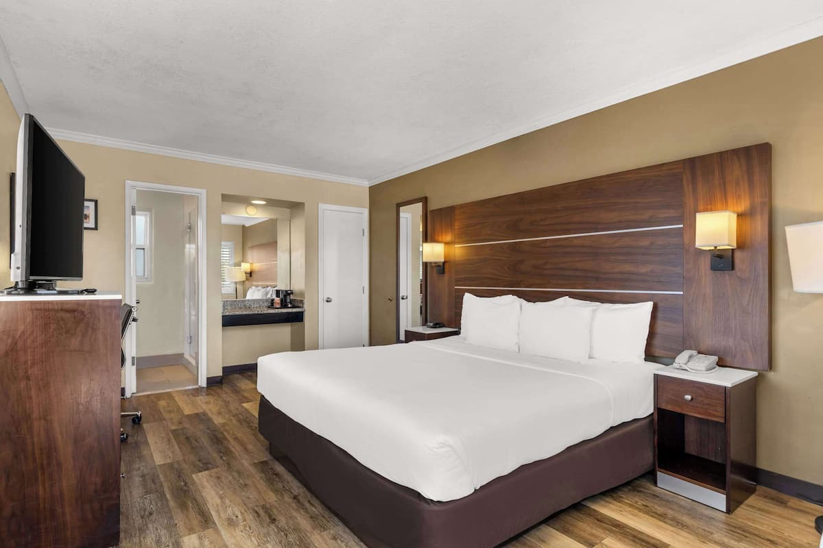 Una habitación de hotel con una cama grande, un cabecero de madera, una mesita de noche con una lámpara, un escritorio con teléfono y un espejo.