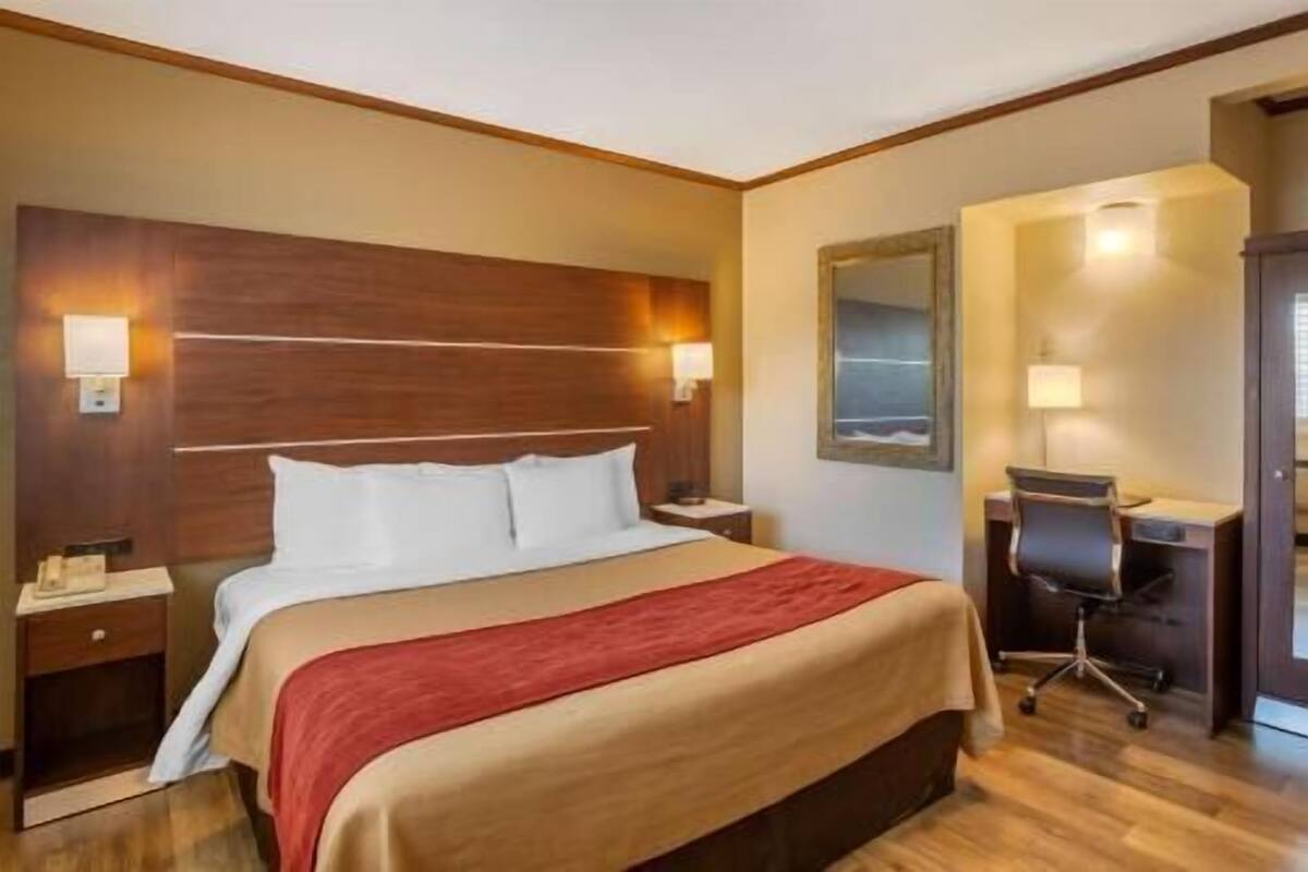 Una habitación de hotel con cabecera de madera, cama con ropa de cama blanca, escritorio con silla y espejo en la pared.