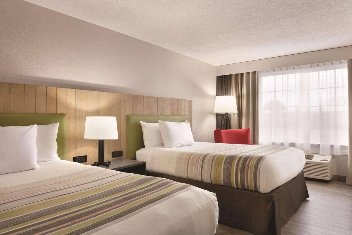 A hotel room with two beds, a wooden headboard, a red chair, a lamp, and a large window with curtains.