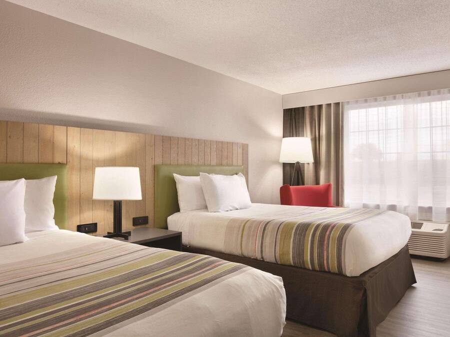 Habitación de hotel con dos camas, cabecero de madera, una silla roja, una lámpara y un ventanal grande con cortinas.