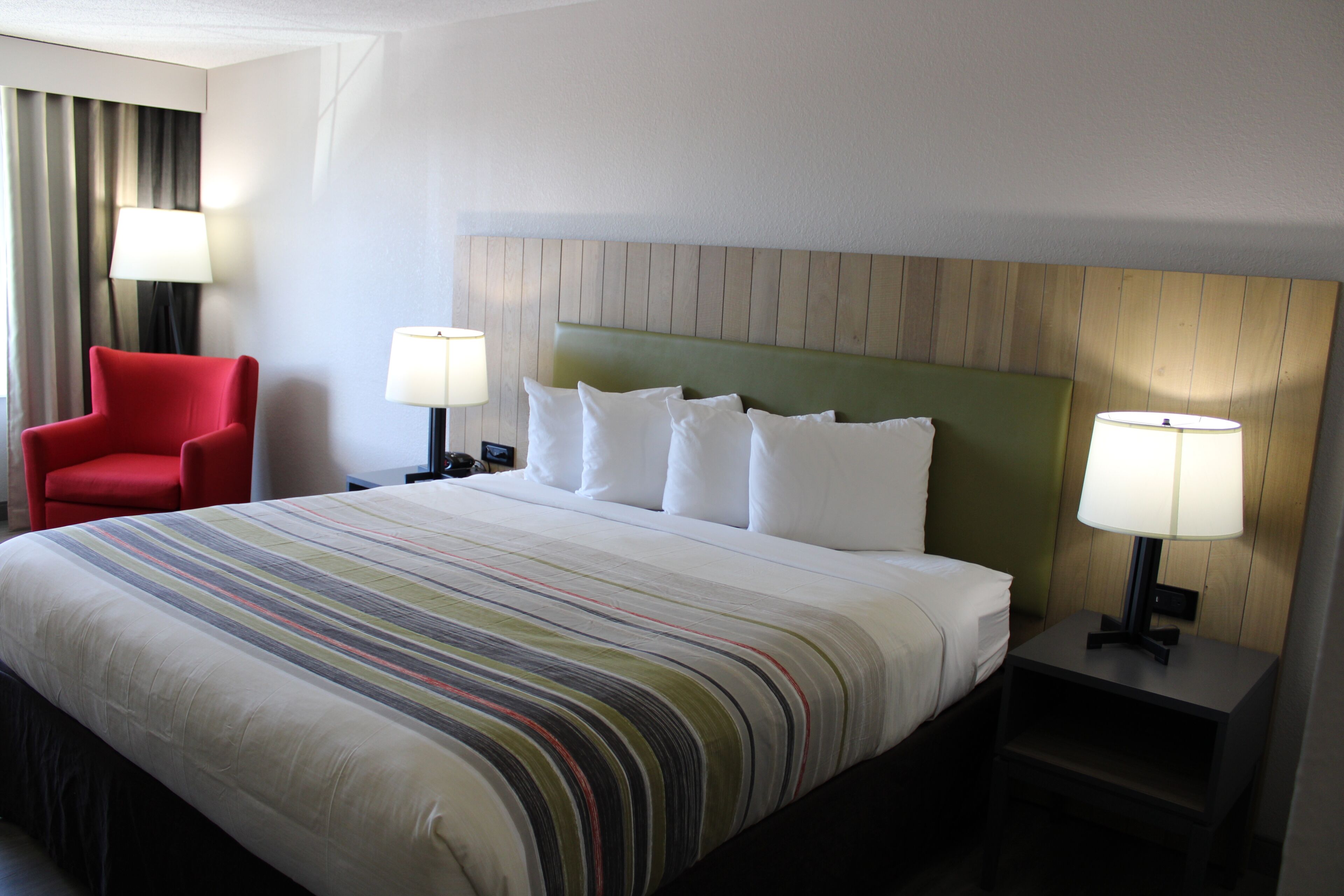 A hotel room with a large bed, two bedside tables with lamps, a red armchair, and a headboard with a wooden panel design.