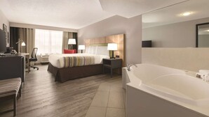 A hotel room with a large bed, a desk with a chair, a desk lamp, a bedside table with a lamp, a large mirror, and a bathtub. - Country Inn & Suites by Radisson, London, KY (London)